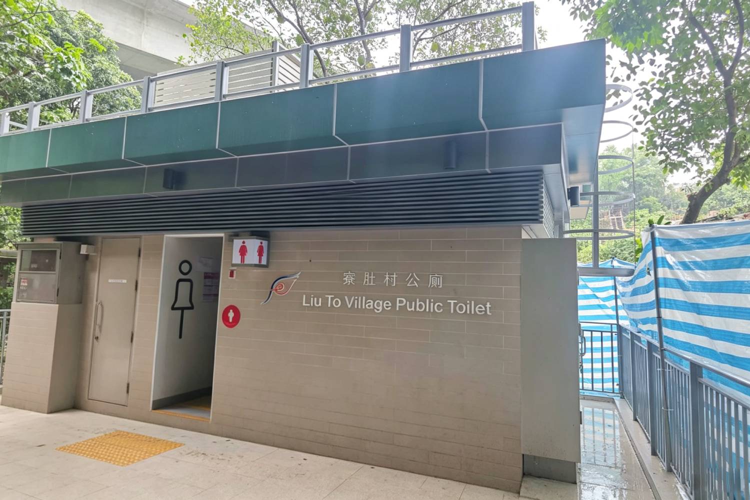 寮肚村公廁外牆鋁合金百葉及鋁窗，香港市政公廁建築外觀，深綠色鋁板頂部結構，橫向鋁通風百葉，淺色牆磚搭配鋁合金門窗，印有「寮肚村公廁 Liu To Village Public Toilet」中英標示，戶外市政建築實拍
