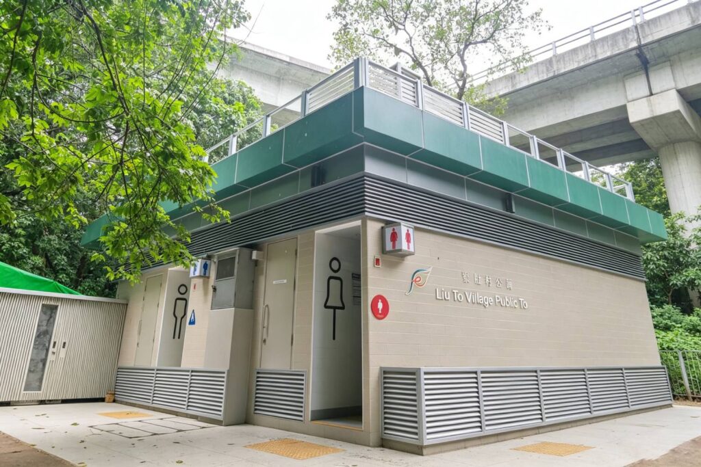 知識庫 寮肚村公廁外牆鋁合金百葉及鋁窗,香港市政公廁建築外觀,深綠色鋁板頂部結構,橫向鋁通風百葉,淺色牆磚搭配鋁合金門窗,印有「寮肚村公廁 Liu To Village Public Toilet」中英標示,戶外市政建築實拍
