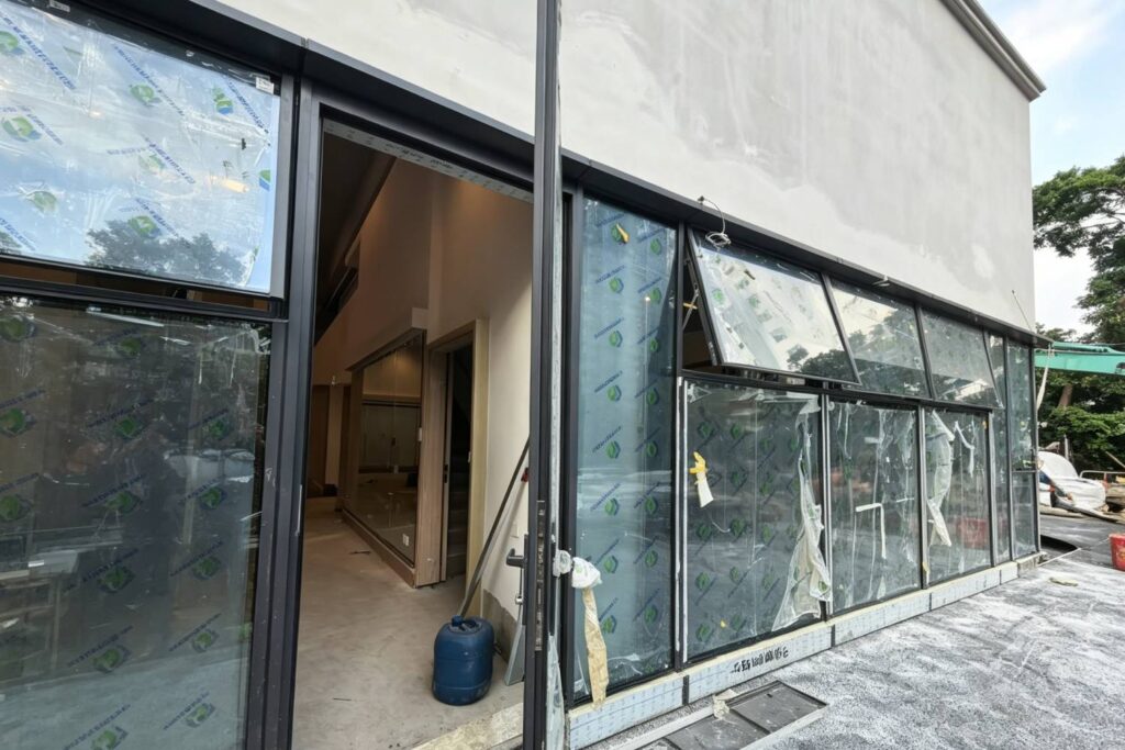 知識庫 黃大仙龍翔道生活萎術中心外牆施工現場,展示新安裝的黑色鋁合金邊框玻璃牆,玻璃表面貼有綠色保護膜,門口敞開可見室內裝修狀況。