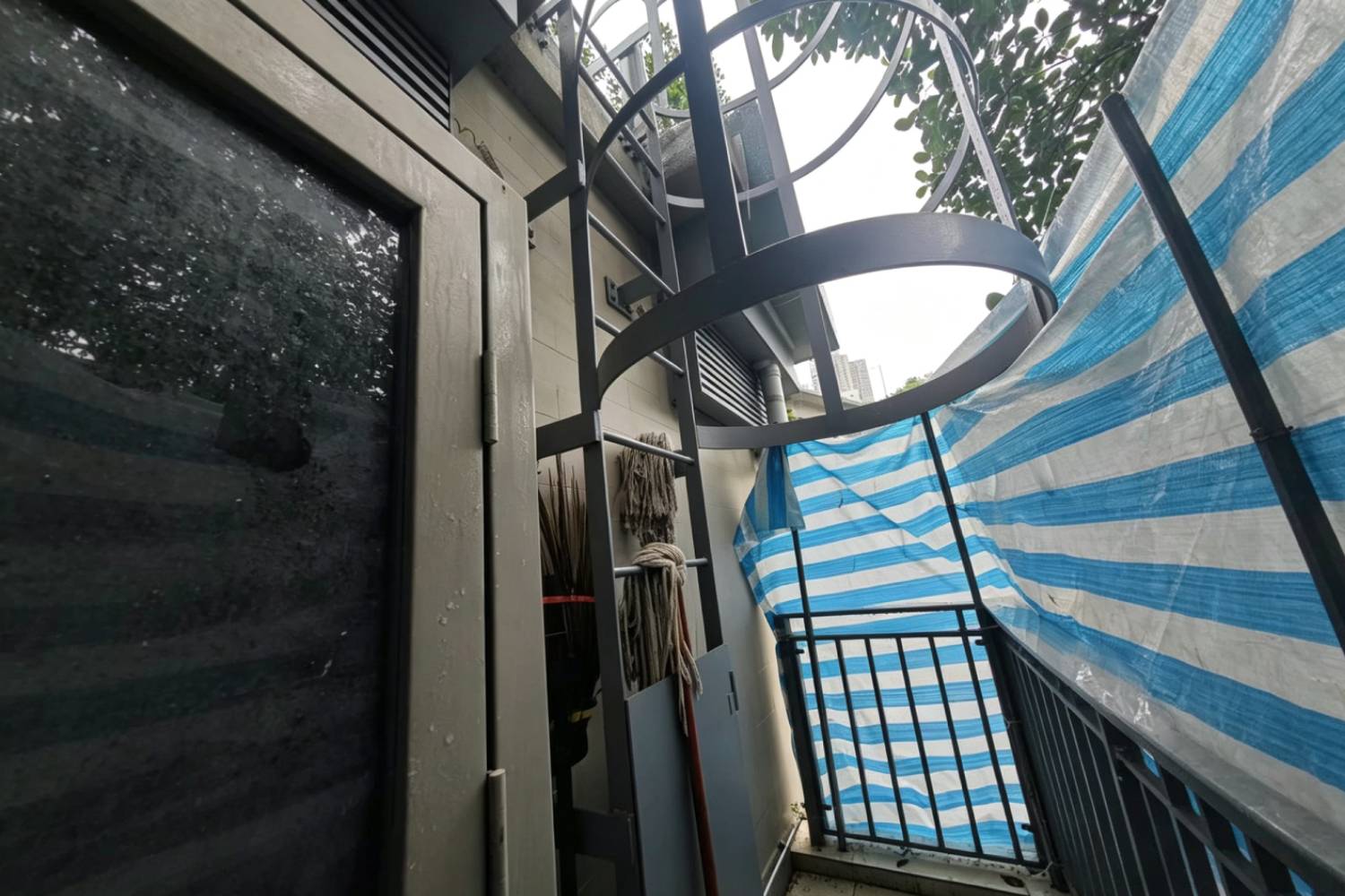 寮肚村公廁戶外貓梯，香港市政建築外牆金屬逃生爬梯，圓形護籠安全梯，灰色金屬結構，旁邊設清潔工具存放區，藍白帆布圍欄，市政工程維修爬梯實拍