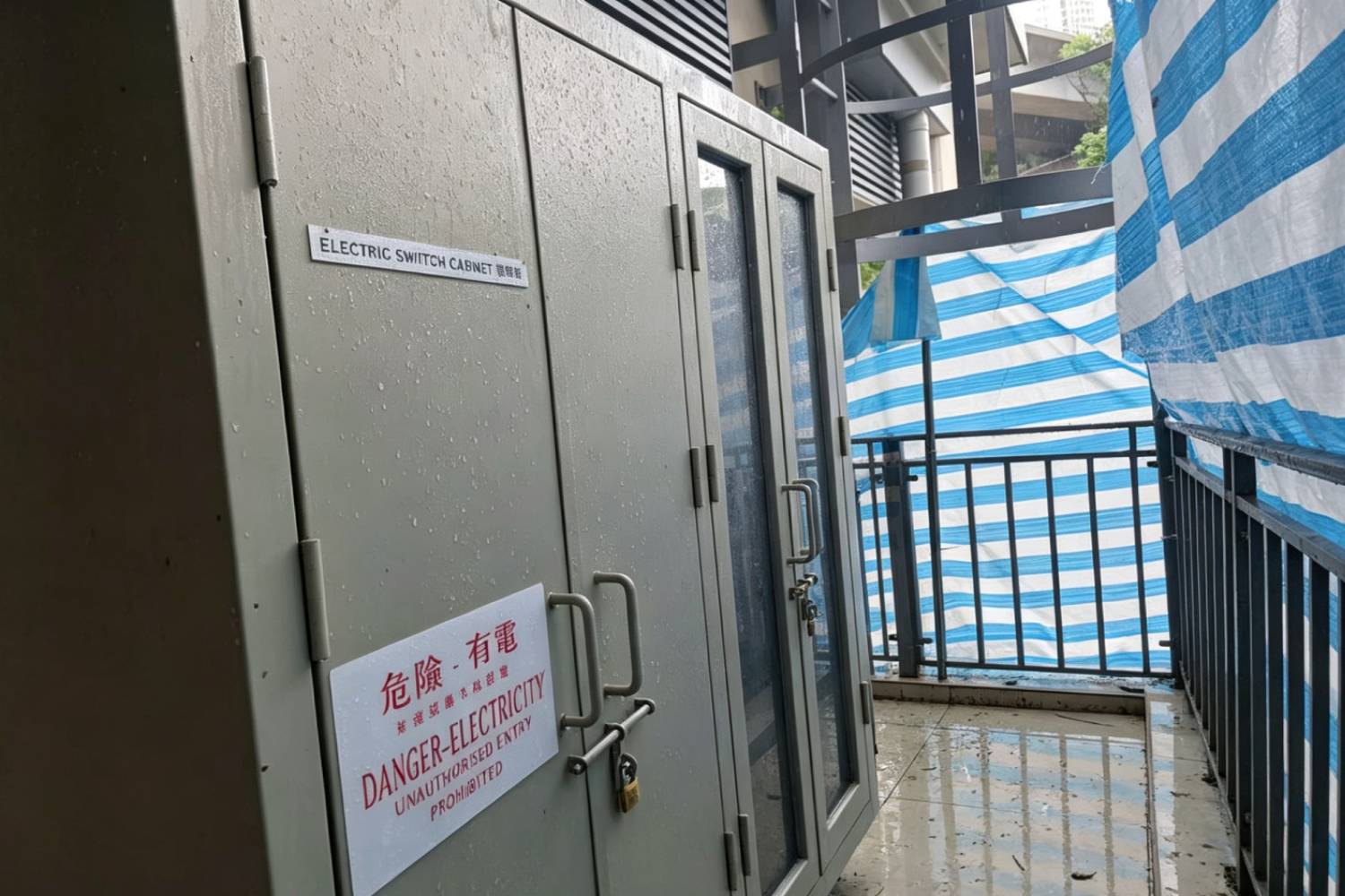 寮肚村公廁戶外不鏽鋼電箱櫃，ELECTRIC SWITCH CABINET 電掣櫃，灰色戶外電力配電櫃，貼有中英「危險 - 有電」警告標籤，配門鎖與金屬把手，市政公廁戶外電力櫃實拍，背景可見藍白帆布圍欄與貓梯結構