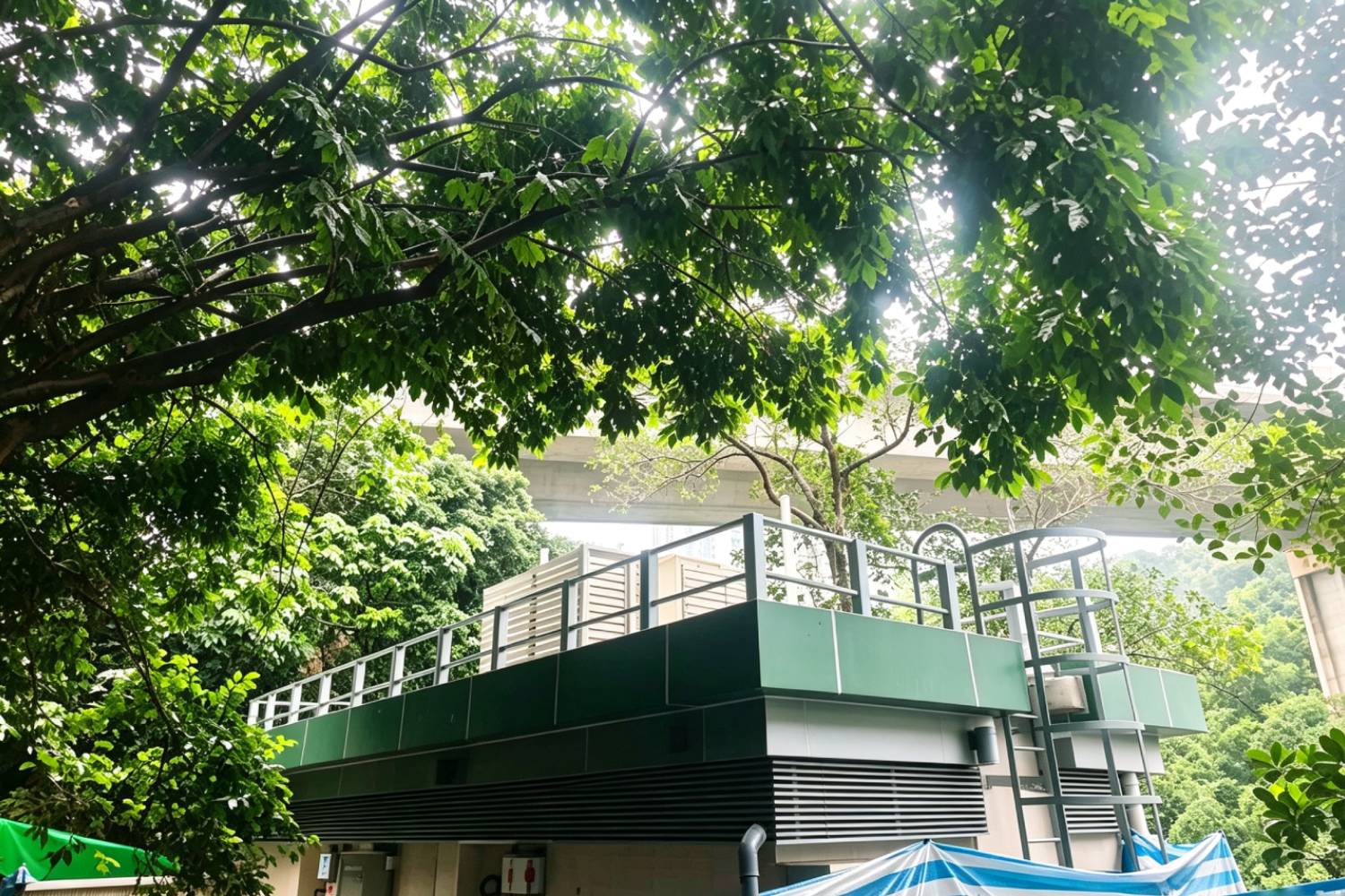 寮肚村公廁全景，鋁合金百葉、屋頂鋁圍欄、不鏽鋼貓梯及不鏽鋼設備箱，香港市政公廁戶外金屬建材，綠色鋁板頂蓋，背景樹木與高架橋，市政工程建築外觀實拍