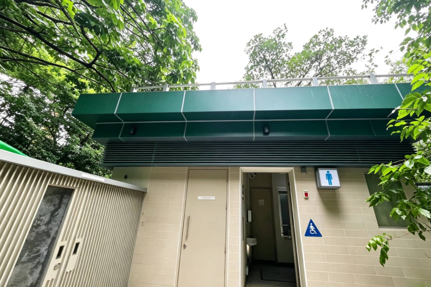 寮肚村公廁外牆鋁合金百葉、鋁合金門窗、屋頂鋁圍欄及側邊不鏽鋼箱，香港市政公廁建築外觀，深綠鋁板頂蓋，橫向通風百葉，淺色牆磚，男廁入口標示，周圍綠樹，市政工程鋁建材與不鏽鋼設備實拍